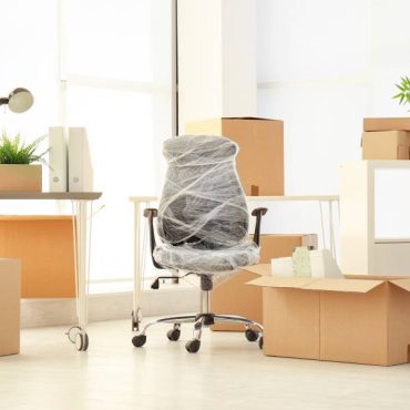 Several stacks of closed cardboard boxes are nestled between office furniture, including plastic-wrapped office chairs.