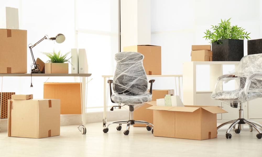 Several stacks of closed cardboard boxes are nestled between office furniture, including plastic-wrapped office chairs.