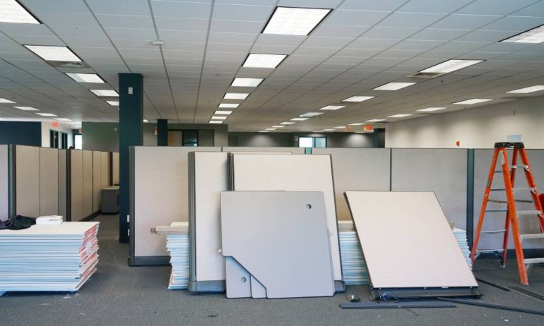 An office undergoing renovations with several stacks of drywall on the ground and an orange ladder in the corner.