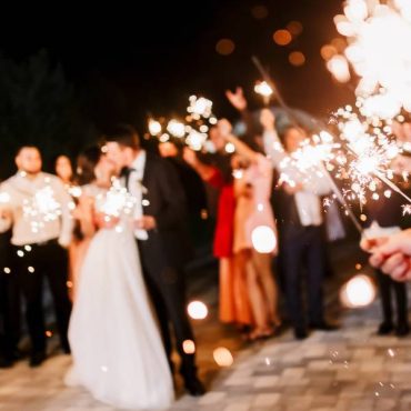 A newlywed couple at their wedding reception enjoying a kiss, while their guests light and hold sparklers around them.