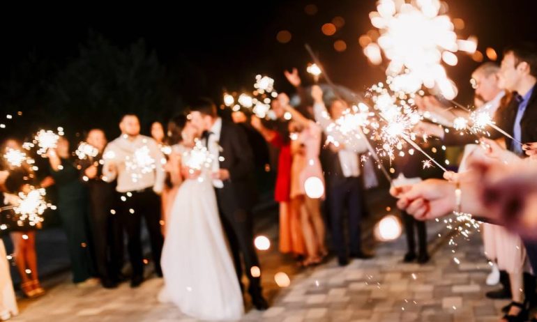 A newlywed couple at their wedding reception enjoying a kiss, while their guests light and hold sparklers around them.