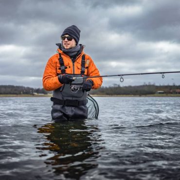 A fisherman in an orange jacket and gray waders stands in the middle of a lake with a fishing pole and net.