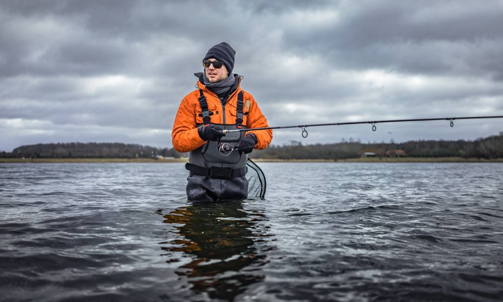 A fisherman in an orange jacket and gray waders stands in the middle of a lake with a fishing pole and net.