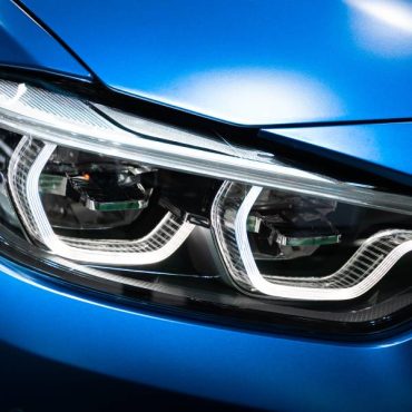 A close view of the activated headlights of a new blue car. The exterior of the vehicle reflects the lights above.