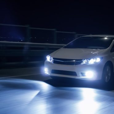 A white car driving in the night with LED headlights turned on. There is a guardrail and fence on the side of the road.