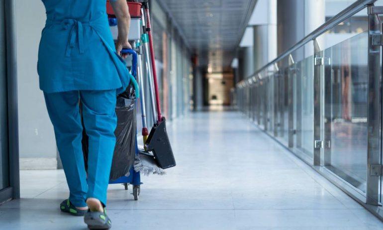 A worker wearing blue scrubs pushing a cleaning cart down a corridor with a glass railing in a commercial building.